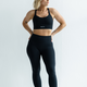 A woman wearing a black cross back sports bra and matching black leggings, standing with one hand on her hip.
